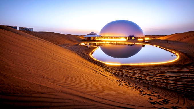 图片展示了沙漠中的巨大圆形建筑，倒映在水面上，四周是沙丘，远处有房屋，景色宁静而美丽。